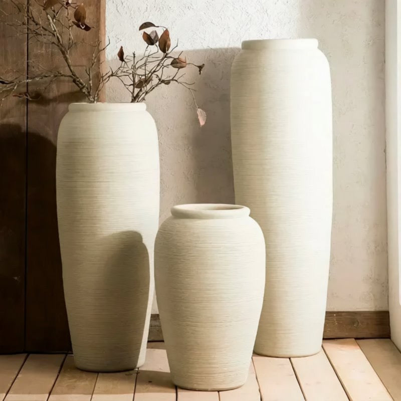 Three tall white vases with a natural plant arrangement in them, placed on a wooden floor against a white wall.